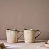 Malia Mug - Cream Set of 2 by Nkuku, showing two 380ml cream stoneware mugs with organic shapes, soft reactive glaze and hand-painted rim detail.