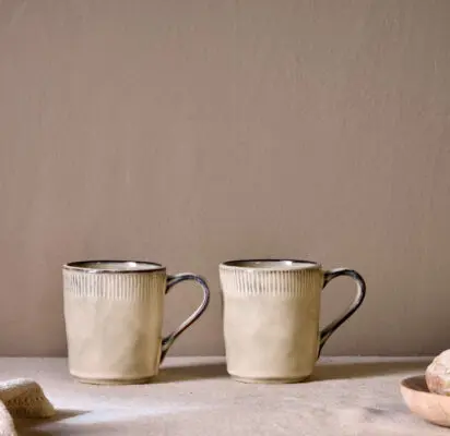 Malia Mug - Cream Set of 2 by Nkuku, showing two 380ml cream stoneware mugs with organic shapes, soft reactive glaze and hand-painted rim detail.