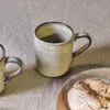 Malia Mug - Cream Set of 2 by Nkuku, showing two 380ml cream stoneware mugs with organic shapes, soft reactive glaze and hand-painted rim detail.