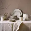 Malia stoneware collection by Nkuku, showing coordinated cream plates, bowls and serving jugs with a soft reactive glaze, fine hand-painted line detail and relaxed organic shapes, styled as a rustic tableware range.