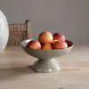Valinha Earthenware Footed Bowl - Grey by Nkuku styled on a table, showing the raised footed base, shallow serving shape and rustic grey earthenware finish.