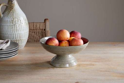 Valinha Earthenware Footed Bowl - Grey by Nkuku styled on a table, showing the raised footed base, shallow serving shape and rustic grey earthenware finish.