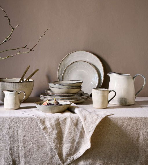 Malia stoneware collection by Nkuku, showing coordinated cream plates, bowls and serving jugs with a soft reactive glaze, fine hand-painted line detail and relaxed organic shapes, styled as a rustic tableware range.