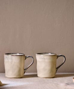Malia Mug - Cream Set of 2 by Nkuku, showing two 380ml cream stoneware mugs with organic shapes, soft reactive glaze and hand-painted rim detail.