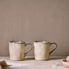 Malia Mug - Cream Set of 2 by Nkuku, showing two 380ml cream stoneware mugs with organic shapes, soft reactive glaze and hand-painted rim detail.