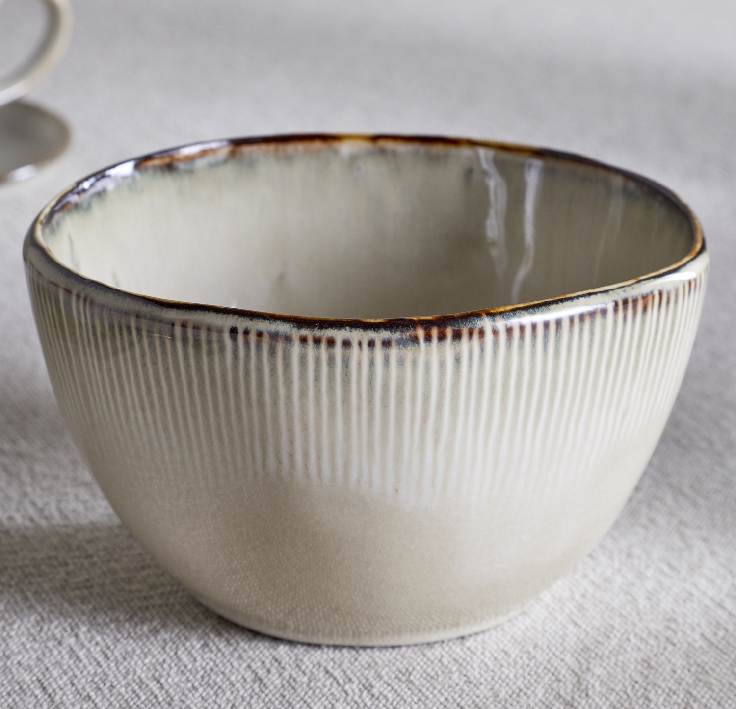 Nkuku Malia Breakfast Bowl in Cream, featuring a softly curved shape, fine vertical ribbed detailing, a warm cream glaze and a contrasting hand-finished brown rim.